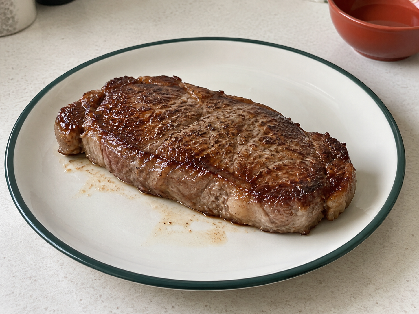 An intact steak with irregular brown surface patches and browned sides rests on a green-rimmed plate with a little juice.